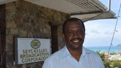 A man stands in front of a building with a sign reading "Seychelles Broadcasting Corporation," with a coastal view in the background.