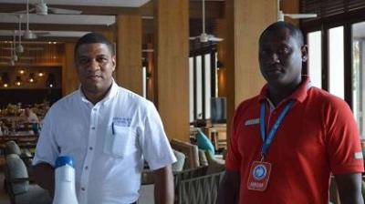 Two men standing indoors, one in a white shirt holding a megaphone, the other in a red shirt with a badge, in a spacious room with wooden pillars and ceiling fans.