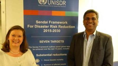 Two people stand smiling in front of a banner for the UNISDR Sendai Framework for Disaster Risk Reduction 2015-2030, outlining seven global targets.