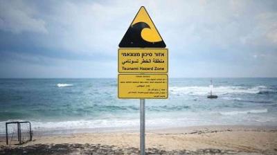 A triangular sign on a beach reads "Tsunami Hazard Zone" in multiple languages, warning of tsunami risk near the ocean.
