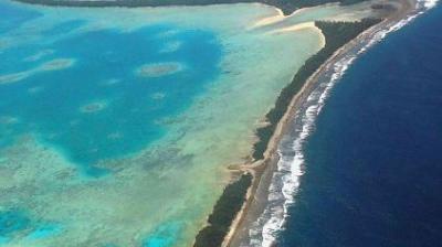 Aerial view of an island with a turquoise lagoon on one side and deep blue ocean on the other, separated by a narrow strip of land with patches of greenery and sandy areas.