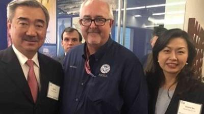Three people pose for a photo at an indoor event. Two men are wearing suits, and the man in the center is wearing a FEMA shirt. A woman stands to the right, smiling.