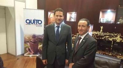 Two men in suits stand in an office with a "Quito" banner in the background. One man is smiling, and the office features dark wood furniture.