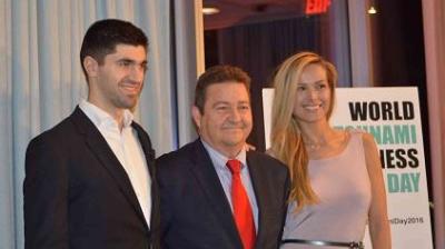 Three people in formal attire stand together, posing for a photo at an event. A sign in the background reads "World Tsunami Awareness Day.