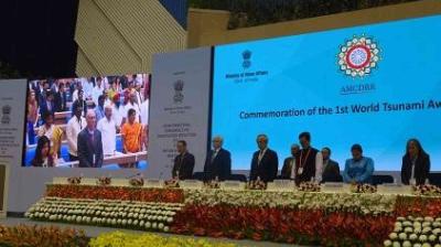 Officials and attendees stand on stage at an event with a screen displaying "Commemoration of the 1st World Tsunami Awareness Day" and floral decorations in front.