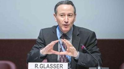 A man in a suit and tie speaks into a microphone at a conference table, seated behind a nameplate labeled "R. Glasser.