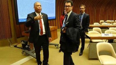 Four people in business attire stand and walk inside a conference room with empty chairs, nameplates, and a large presentation screen in the background.