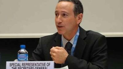 A man in a suit speaks at a podium with a sign reading "Special Representative of the Secretary-General." A bottle of water is placed in front of him.