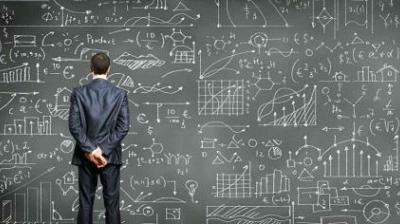 A man in a suit stands facing a large chalkboard filled with complex graphs, charts, and mathematical formulas.