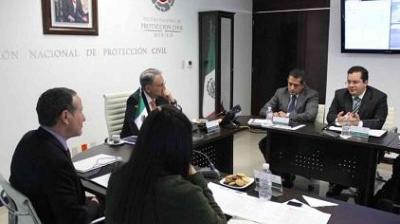 A group of people in a meeting room with folders and laptops on the table. A screen and national emblem are visible on the wall.
