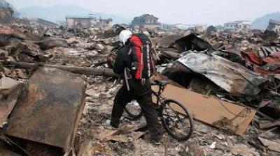 A person wearing a backpack and helmet walks a bicycle through debris and rubble in a devastated area with damaged buildings in the background.