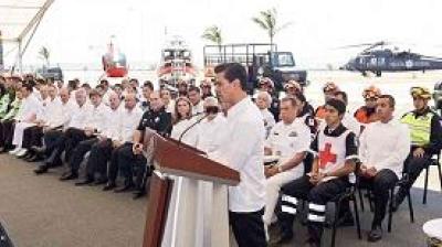 A person speaks at a podium in front of a seated audience, including uniformed personnel and Red Cross members, with emergency vehicles visible in the background.
