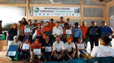A group of people pose indoors holding certificates under a banner that reads “Bienvenidos a Cedeño Comunidad ‘Tsunami Ready’.”.