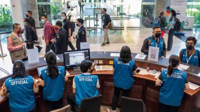 Officials in blue vests assist attendees at a registration desk in a lobby area; people walk and converse nearby.