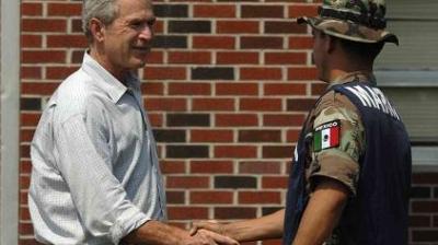 A man in a light shirt shakes hands with a uniformed soldier wearing a helmet and a vest labeled "MARINA" with a Mexican flag patch, in front of a brick wall.