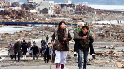 A group of people walk through a devastated area filled with debris; one woman carries an elderly person on her back.