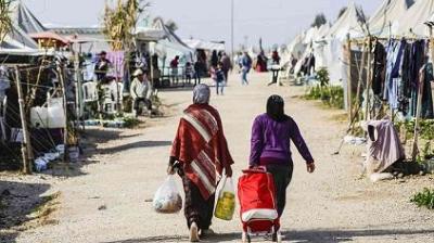 Two people walk down a dirt path in a tented area, carrying goods and a cart, with other people and tents visible along the sides.
