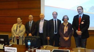 Six people in business attire stand in a row at a conference room, with nameplates and microphones visible on the table in front of them.