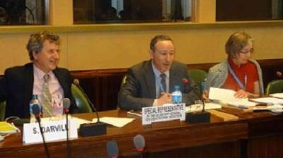 Three people sit at a conference table with microphones in front of them. Two men and a woman are engaged in discussion, with name placards and documents on the table.