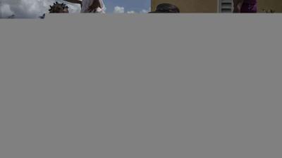 A child stands on a ledge reaching toward the sky, while another child watches from below. The background includes a yellow building and blue sky with clouds.