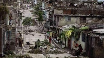 A damaged urban street with debris, fallen plants, and partially destroyed buildings. People are walking and standing amidst the rubble.