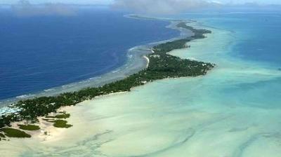 Aerial view of a narrow tropical island surrounded by turquoise shallows and deep blue ocean, with scattered vegetation and sandy beaches.