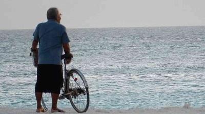 An older person stands barefoot on the beach holding a bicycle, looking out at the calm sea under a clear sky.