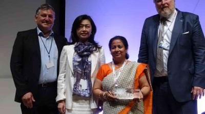 Four people pose for a photo at an event; one person, dressed in an orange saree, holds a glass award.