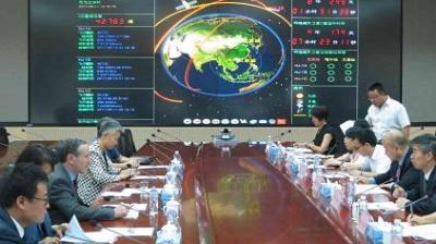 People seated on both sides of a conference table with documents, in front of a large digital screen displaying satellite data and a map of Asia.