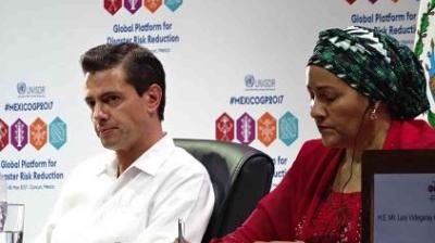 Two individuals sit at a panel, with a backdrop displaying "Global Platform for Disaster Risk Reduction" and various logos. One is writing; the other listens attentively.