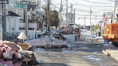 A residential street is lined with debris, damaged furniture, and trash piles, with utility poles and houses visible on both sides. People are in the background near an orange dumpster.