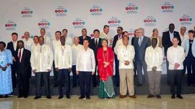 A group of delegates in formal attire pose for a photo in front of a conference backdrop with logos and event branding.