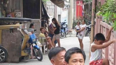 People are gathered in a narrow street. A child climbs a wall, while others sit or stand nearby. Motorcycles and a tricycle are parked along the street.