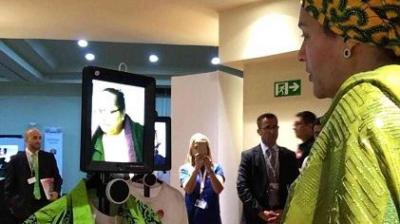 A person in green attire speaks to another person on a screen through a video call using a mobile device mounted on a rolling stand. Several people are standing in the background.