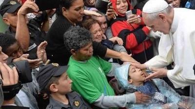 Pope Francis touches the face of a child in a wheelchair, surrounded by a crowd of people, some taking photos and police officers present.