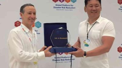 Two men stand together as one presents an award plaque to the other at an event for disaster risk reduction. Both are smiling and wearing white shirts.