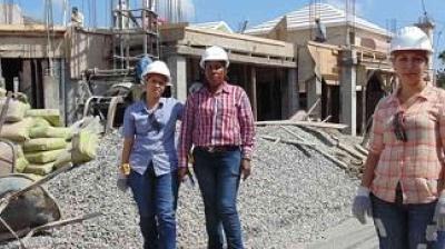 Three construction workers wearing hard hats stand in front of a building under construction, with scaffolding, gravel, and cement bags visible.