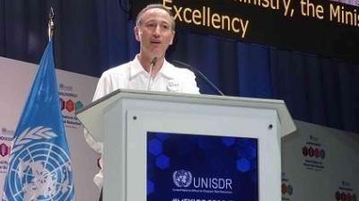 A man stands at a podium with a UNISDR logo, speaking at a conference; a United Nations flag is visible beside him.