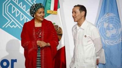 Two people stand in front of Mexican and UN flags, exchanging glances. One wears a red outfit and headscarf, the other a white shirt. They are in a setting with a blue and white background.