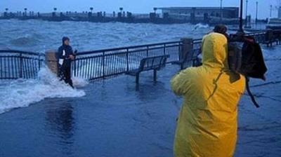 A person in yellow rain gear films a reporter standing by a flooded waterfront with waves crashing over a railing during stormy weather.