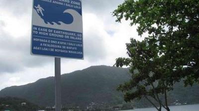 Tsunami warning sign by a coastline with mountains in the background, advising to move to high ground or inland in case of an earthquake.