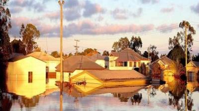 Residential neighborhood with houses partially submerged in floodwater, reflecting the buildings and sky, with trees and power lines in the background.