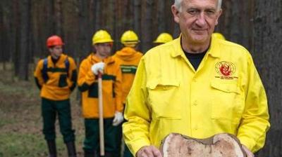 A man in a yellow uniform holds a slice of tree trunk, while four people in safety gear stand in the background in a forest.