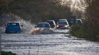 Cars driving through a flooded road with water splashing around. Trees line both sides of the road.