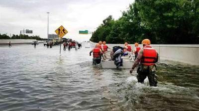 People wearing life vests wade through a flooded highway, some pulling a small boat with belongings, while others walk further ahead.