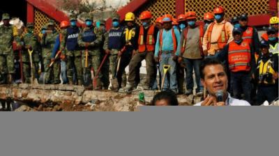 A group of rescue workers and officials in safety gear stand in a line at a disaster site while a man speaks into a microphone in the foreground.