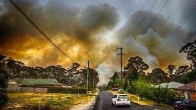A rural road with houses is overshadowed by thick smoke from a large wildfire in the background, filling the sky with clouds of smoke.
