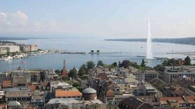 Aerial view of a city with buildings in the foreground, a large lake with a prominent water fountain in the center, and distant mountains under a clear sky.