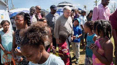 A group of adults and children gather outdoors near tents on a sunny day, some standing and some talking among themselves.