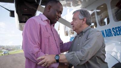 Two men are standing on a tarmac in front of a small aircraft, shaking hands and engaging in conversation.